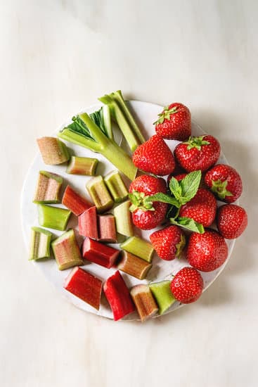 Fresh organic garden strawberries and cutting rhubarb stems on ceramic board over white marble background. Flat lay, space. Ingredients for summer lemonade, jam or cake